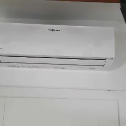 Biały klimatyzator Viessmann DC Inverter zamontowany na białej ścianie, oświetlony przez dwa reflektory. Widoczny fragment drzwi.