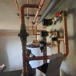 Wailet Grzegorz Chludziński - Instalacja miedzianych rur grzewczych w surowym wnętrzu, widoczna osoba w trakcie montażu, skrzynka z narzędziami obok. Świeżo położone rury w nowym budynku.