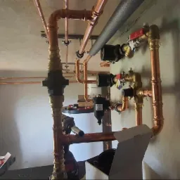 Instalacja miedzianych rur grzewczych w surowym wnętrzu, widoczna osoba w trakcie montażu, skrzynka z narzędziami obok. Świeżo położone rury w nowym budynku.