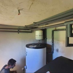 Wailet Grzegorz Chludziński - Złożona instalacja grzewcza w surowym pomieszczeniu z widocznym zasobnikiem ciepłej wody i rurami. Fachowiec przyłącza elementy instalacji.