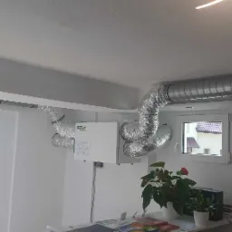 Zamontowana centrala rekuperacyjna WANAS z widocznymi przewodami wentylacyjnymi w pomieszczeniu o białych ścianach i suficie. Widoczne okno i roślina doniczkowa.