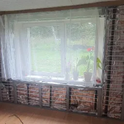 Ściana w trakcie montażu ogrzewania ściennego, widoczne rury grzewcze w metalowej konstrukcji, przygotowane do zabudowy płytami kartonowo-gipsowymi. Na parapecie okiennym stoją doniczki z kwiatami.