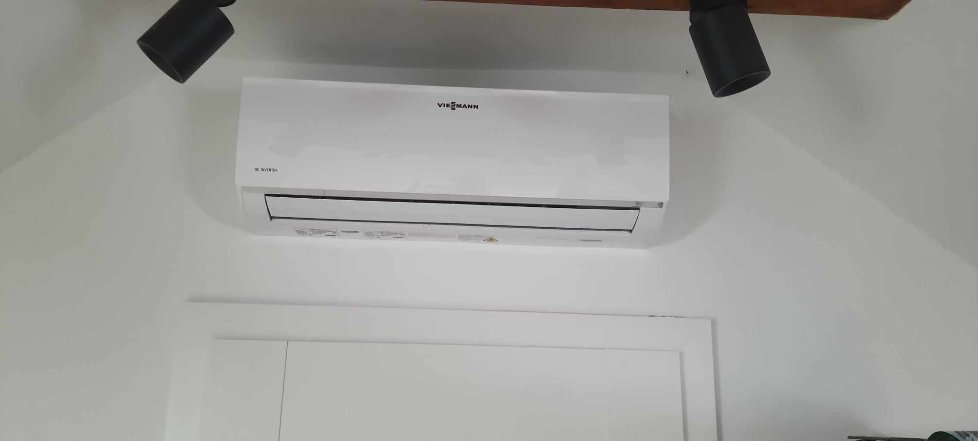 Biały klimatyzator Viessmann DC Inverter zamontowany na białej ścianie, oświetlony dwoma reflektorami. Widok z dołu na urządzenie w minimalistycznym wnętrzu.