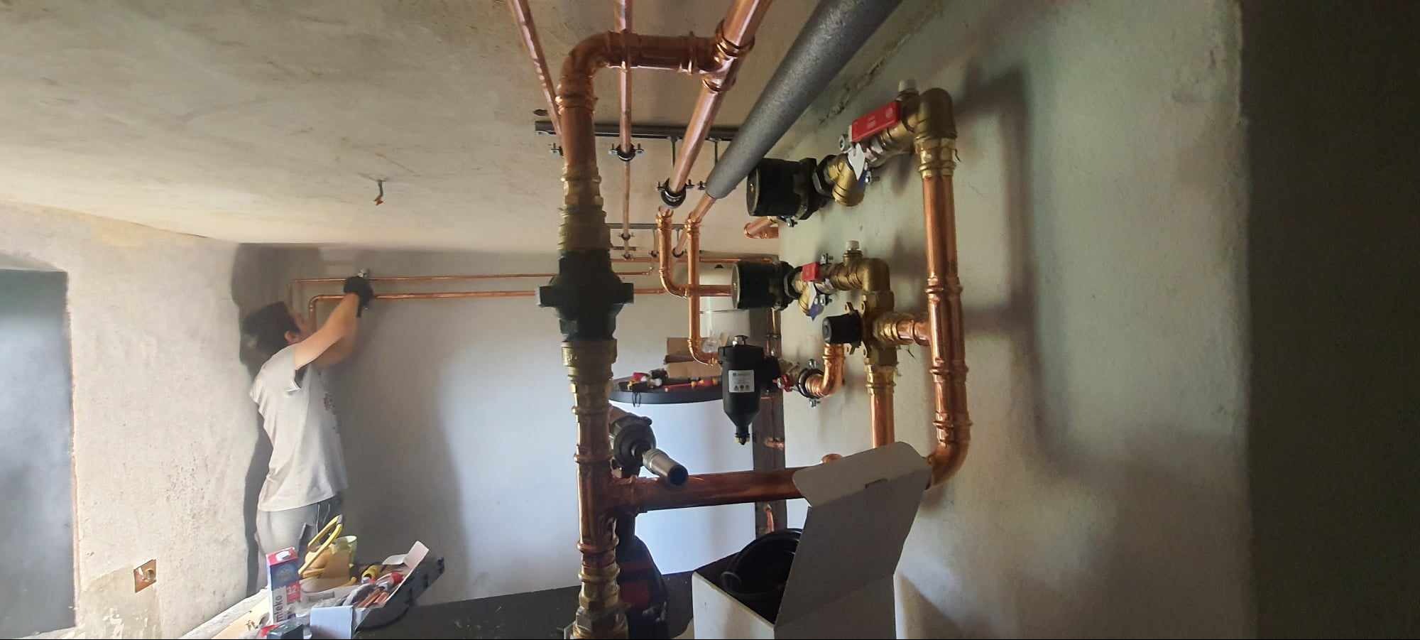 Instalacja miedzianych rur grzewczych w surowym wnętrzu, widoczna osoba w trakcie montażu, skrzynka z narzędziami obok. Świeżo położone rury w nowym budynku.