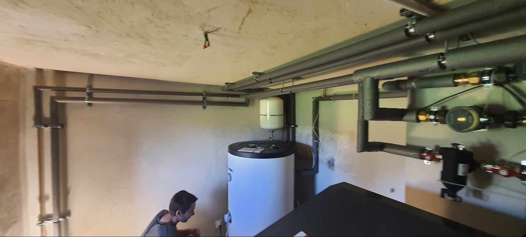 Złożona instalacja grzewcza w surowym pomieszczeniu z widocznym zasobnikiem ciepłej wody i rurami. Fachowiec przyłącza elementy instalacji.