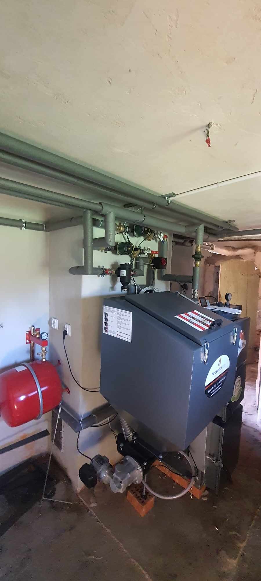 Szary piec z podłączonymi rurami w kotłowni. Widoczny czerwony zbiornik wyrównawczy i instalacja na ścianie. Nierówne ściany i sufit.