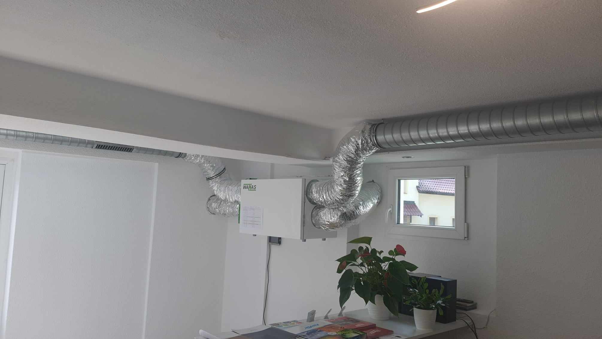 Zamontowana centrala rekuperacyjna WANAS z widocznymi przewodami wentylacyjnymi w pomieszczeniu o białych ścianach i suficie. Widoczne okno i roślina doniczkowa.