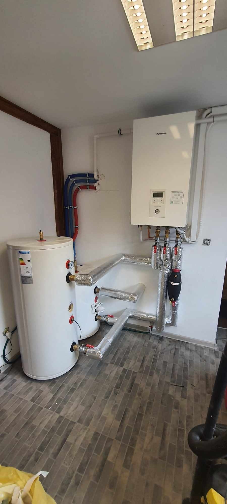 Biały zasobnik ciepłej wody z podłączonym kotłem gazowym Panasonic i rurami w izolacji aluminiowej, na tle białej ściany i szarej podłogi.