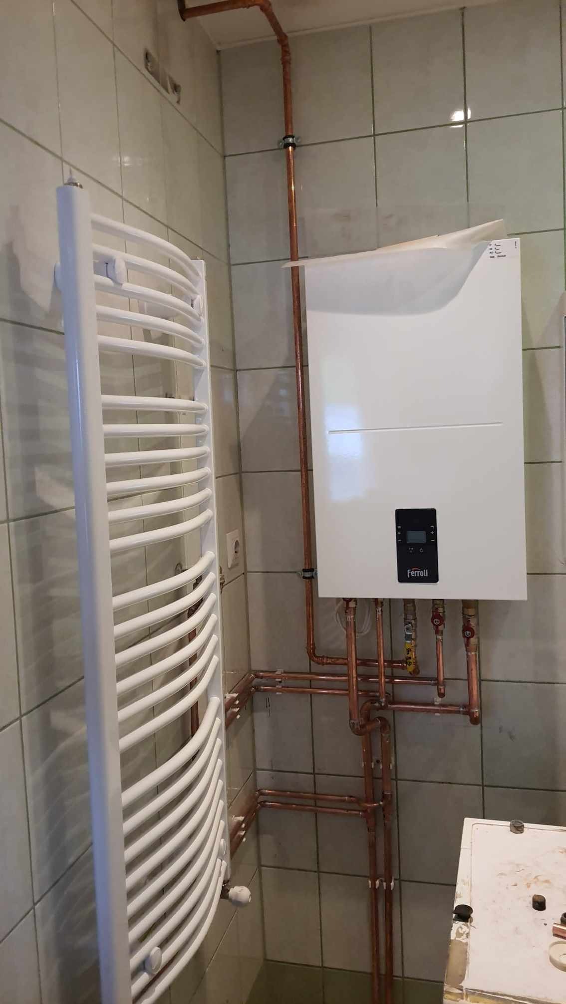 Biały, nowoczesny piec gazowy Ferroli zamontowany w łazience z grzejnikiem drabinkowym i miedzianą instalacją. Widoczne płytki ceramiczne na ścianach.