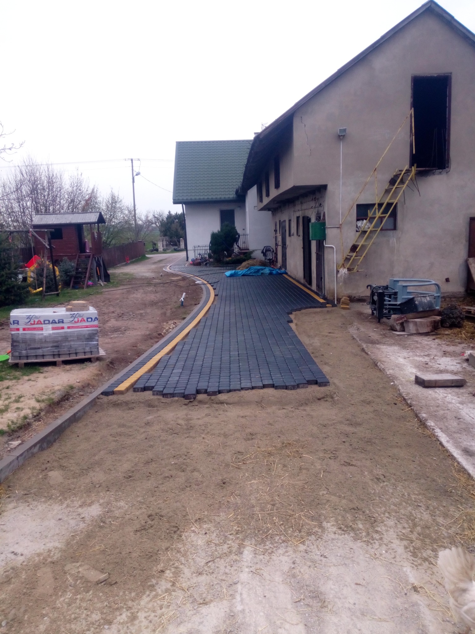 Częściowo ułożona, czarna kostka brukowa z żółtym obrzeżem na posesji w wiejskim otoczeniu, paleta kostki w tle.