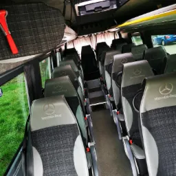 Wnętrze autobusu Mercedes-Benz z rzędami foteli pasażerskich w szaro-czarnej kolorystyce z logo marki na zagłówkach, widoczne stopnie wejściowe i okna z zielonym otoczeniem na zewnątrz.
