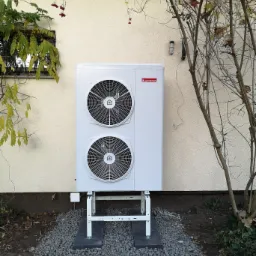 Biała pompa ciepła Ariston zamontowana na zewnątrz budynku z beżową elewacją, widok z dołu, z krzewem i oknem w tle.