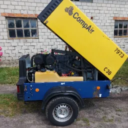 Przenośny kompresor marki CompAir z otwartą żółtą obudową na nieutwardzonym podłożu, widoczny silnik i osprzęt, w tle murowany budynek z oknami i różowe kwiaty.