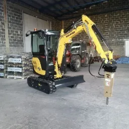 Żółto-czarna mini koparka Yanmar z zamontowanym młotem hydraulicznym, stojąca w hali z betonową podłogą, w tle widoczny ciągnik i palety z materiałami budowlanymi.