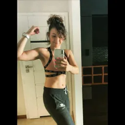 Kobieta w sportowym stroju robi selfie, prezentując napięty biceps. Widoczny zegarek na nadgarstku i sportowa bielizna z logo.