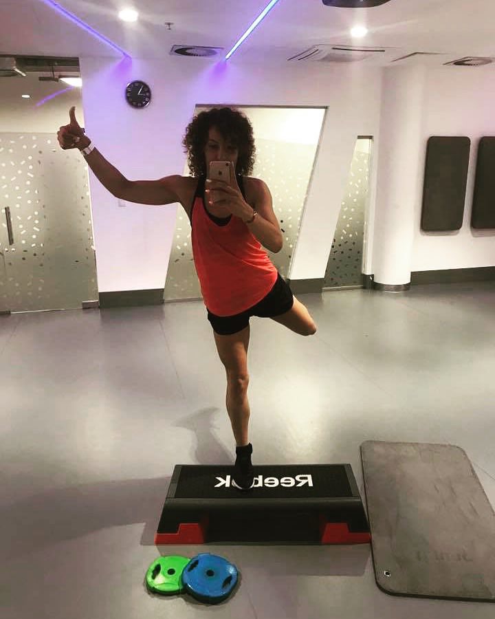 Kobieta robiąca selfie w lustrze, stojąc na jednej nodze na stepie Reebok, z uniesionym kciukiem, w tle widoczna sala gimnastyczna z zegarem.