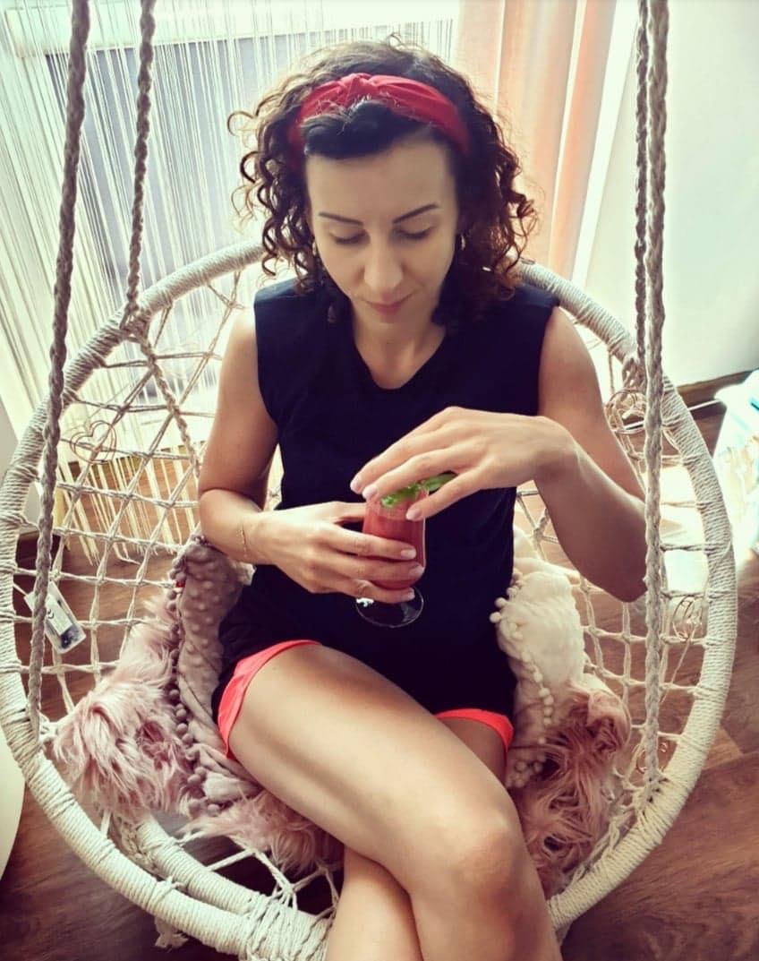 Kobieta z czerwoną opaską na włosach siedzi w wiszącym fotelu, trzyma szklankę z czerwonym koktajlem i listkiem mięty.