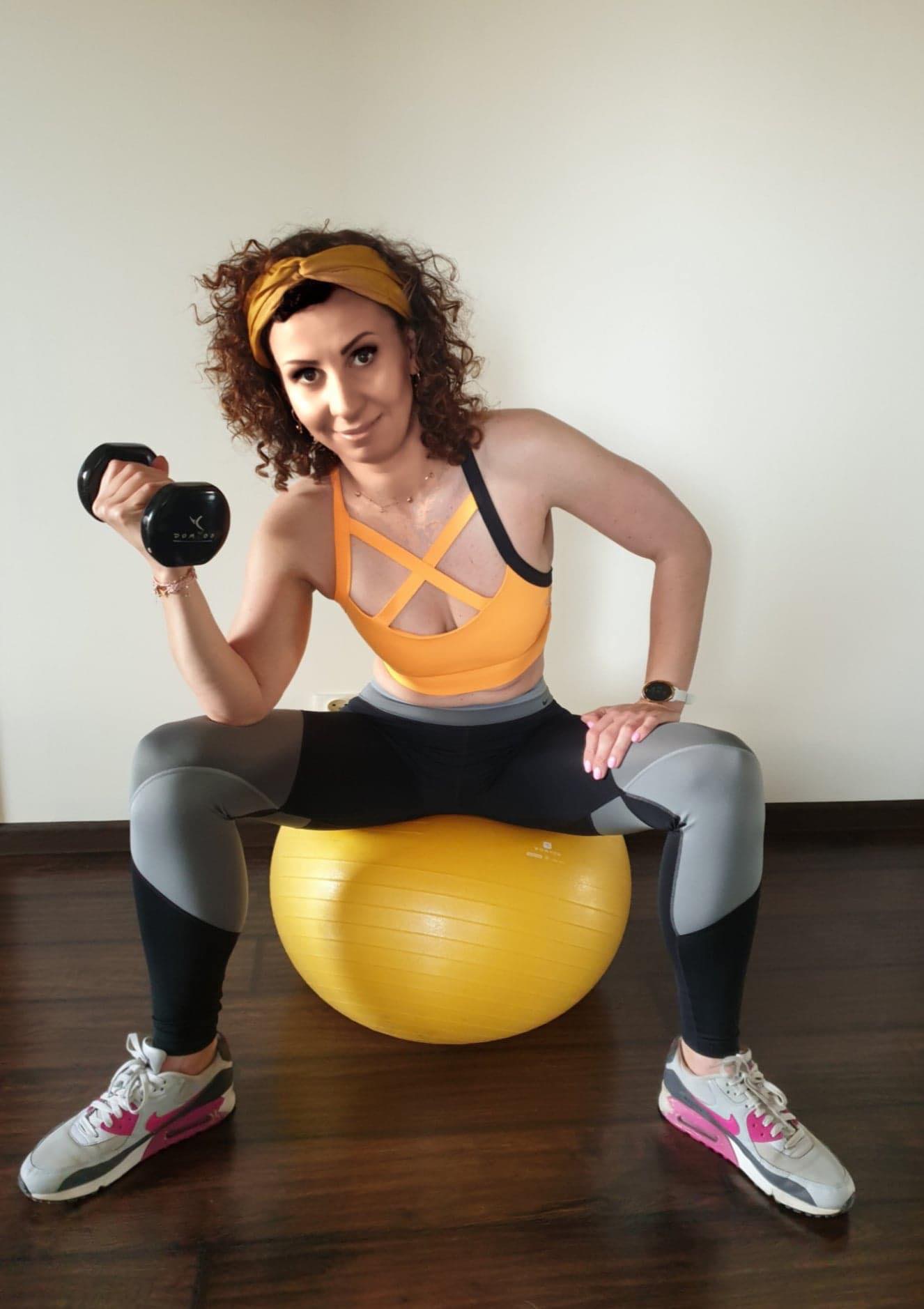 Kobieta w sportowym stroju siedzi na żółtej piłce do ćwiczeń, unosząc hantel w prawej ręce, na tle jasnej ściany i ciemnej podłogi.