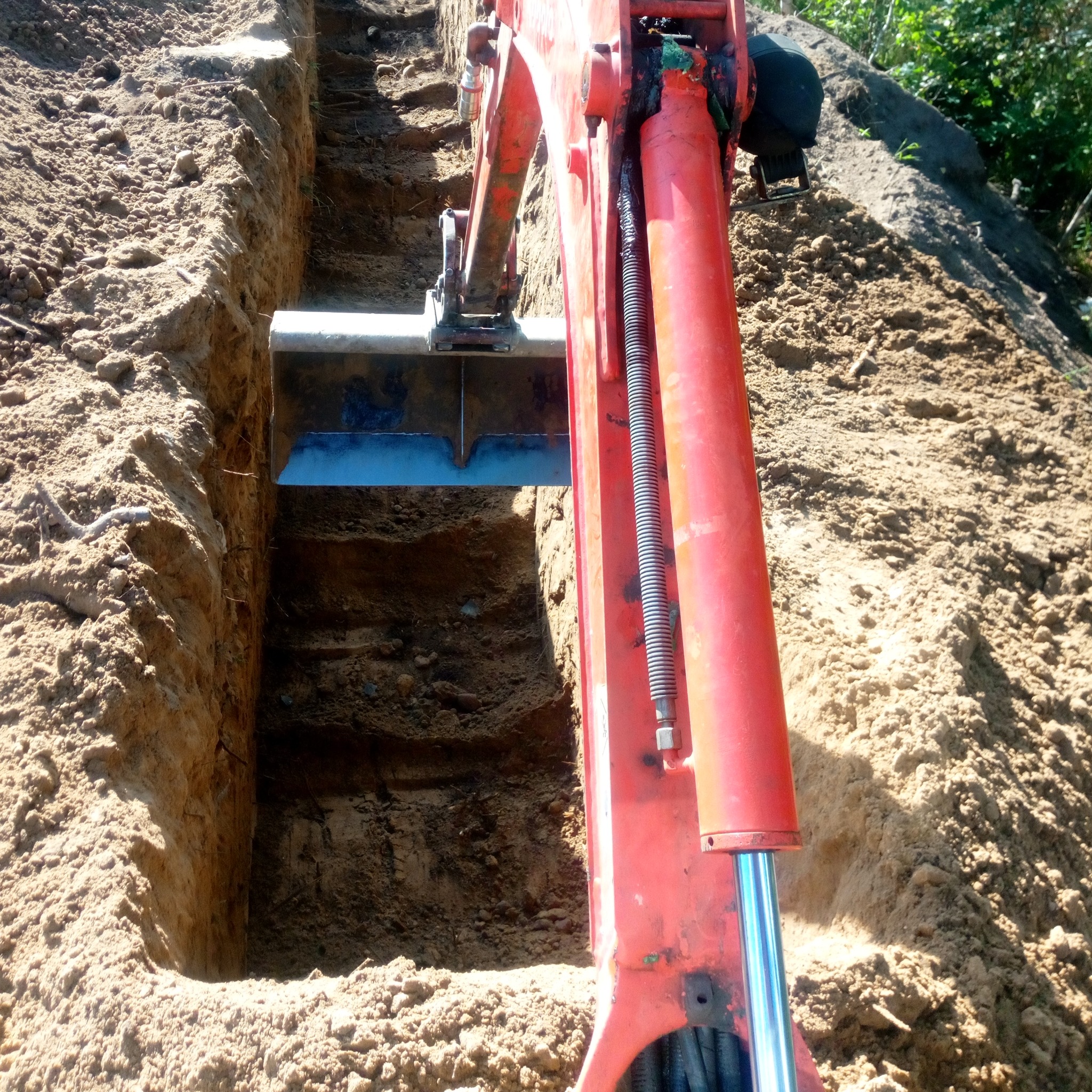 Czerwona koparka w trakcie wykonywania wąskiego wykopu w piaszczystej glebie, widoczne ślady pracy łyżki i nierówne ściany wykopu.