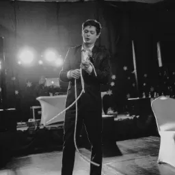 Czarno-białe zdjęcie iluzjonisty w garniturze, trzymającego w dłoniach białą linę na scenie z dekoracjami w tle, podczas występu.