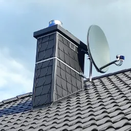 Antena satelitarna zamontowana na dachu obok komina z grafitową dachówką karpiówką, widok z dołu na tle błękitnego nieba.