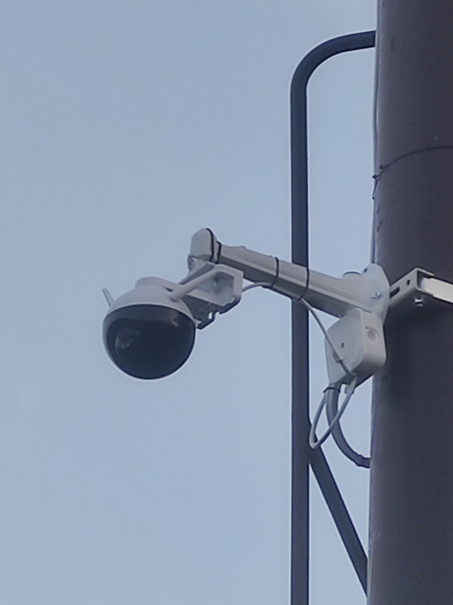 Kamera monitoringu kopułkowa zamontowana na słupie, widoczne przewody i uchwyt montażowy, tło stanowi jasne niebo.