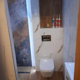 Nowoczesna łazienka z płytkami imitującymi marmur, podwieszana toaleta, drewnopodobna podłoga i wnęka z drewnianą okładziną, na której stoją tuby z silikonem.