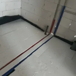 Białe płyty styropianowe na podłodze w trakcie montażu, z widocznymi rurami instalacyjnymi w kolorze niebieskim i czerwonym oraz fragmentem czarnej folii hydroizolacyjnej przy ścianie z białych...