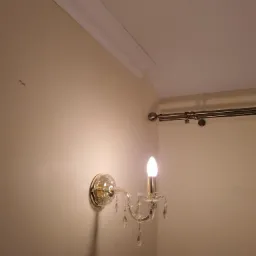 Kryształowy kinkiet z żarówką w kształcie płomienia zamontowany na beżowej ścianie z listwą przysufitową i karniszem