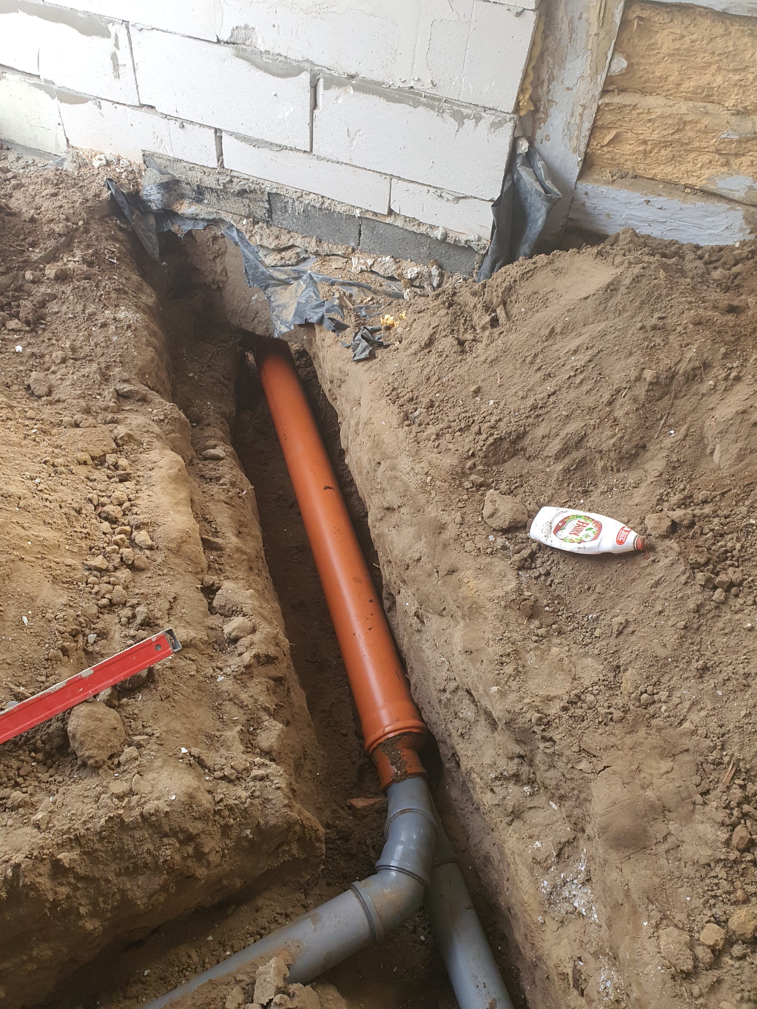 Wykop w ziemi z pomarańczową rurą kanalizacyjną ułożoną wzdłuż ściany fundamentowej z pustaków, widoczny poziomica i butelka płynu do naczyń na ziemi.