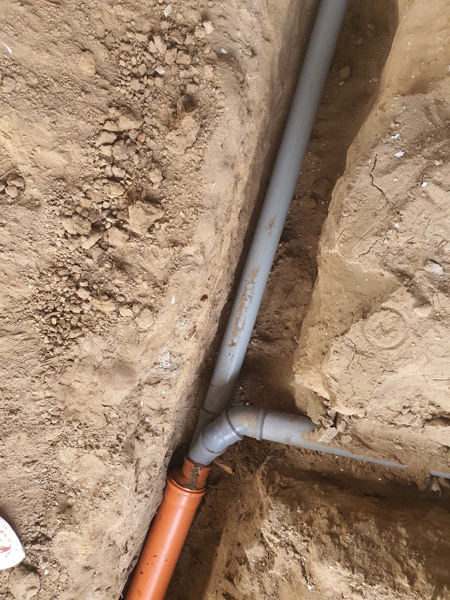 Pomarańczowa i szara rura kanalizacyjna ułożona w wykopie w ziemi, widok z góry.