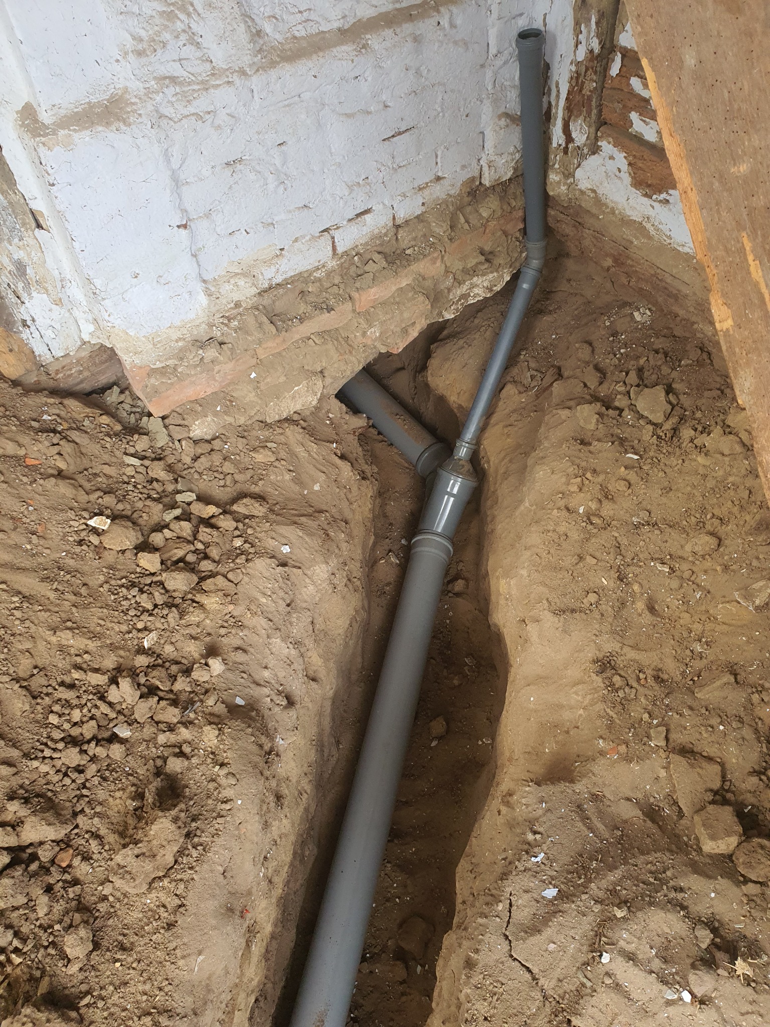 Szare rury kanalizacyjne ułożone w wykopie ziemnym wzdłuż ściany z cegły i fragmentu drewnianej konstrukcji, widoczne złącza i pionowy przebieg rury.