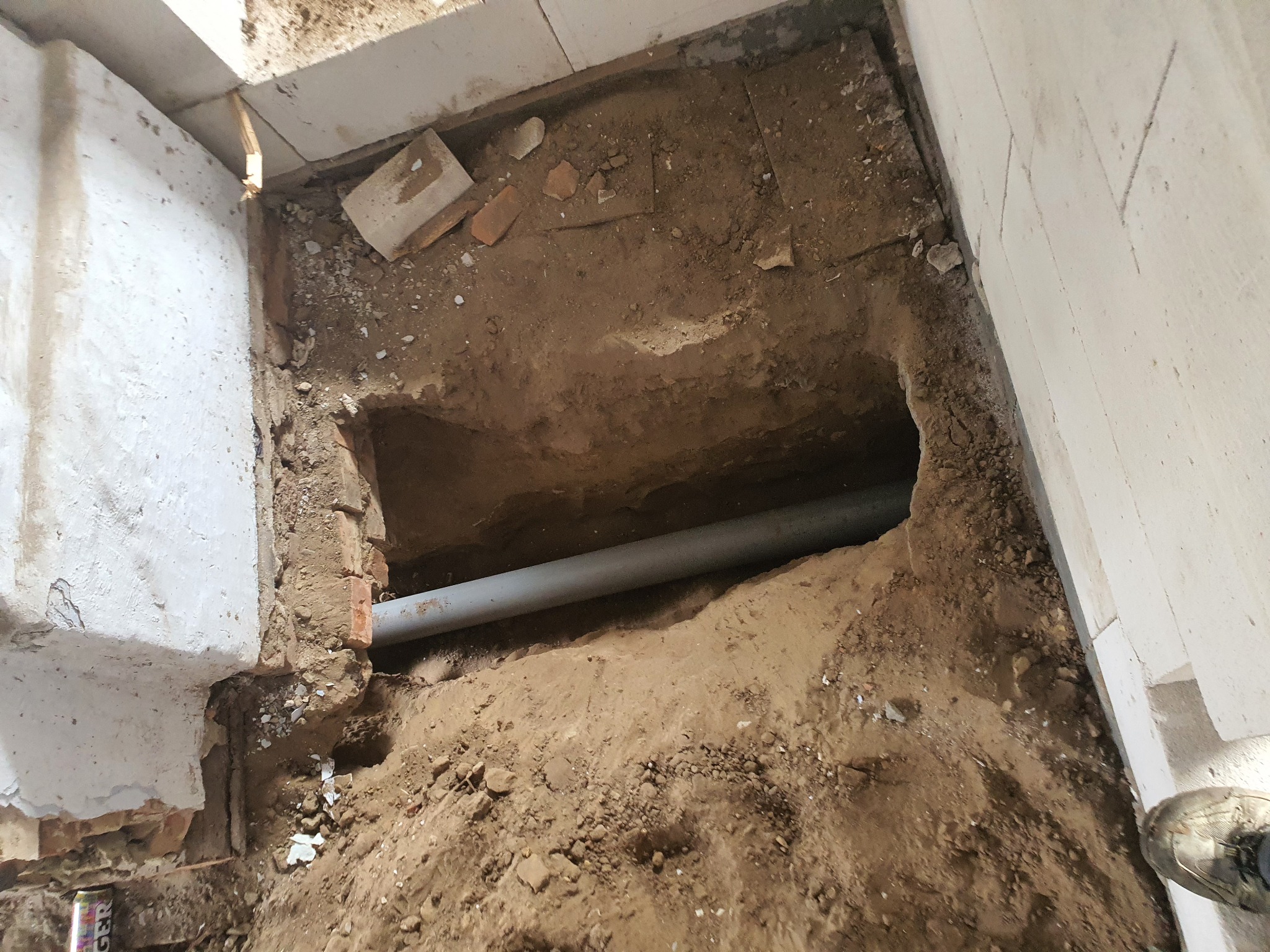 Wykop w ziemi wewnątrz budynku z widoczną rurą kanalizacyjną, fragmentami cegieł i pustaków oraz ścianą z nierówną fakturą.