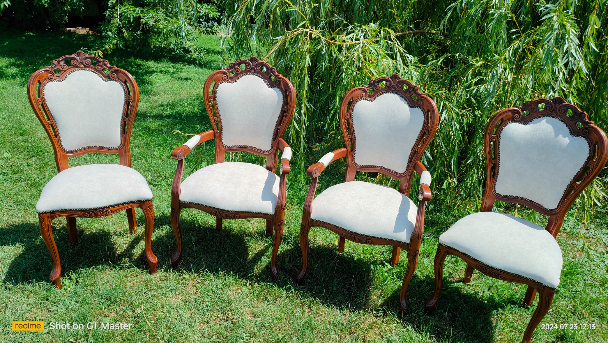 Cztery odnowione krzesła w stylu Ludwika na trawie, z jasną tapicerką i ciemnobrązową, rzeźbioną ramą z dekoracyjnymi ćwiekami, ustawione na tle zieleni drzew.