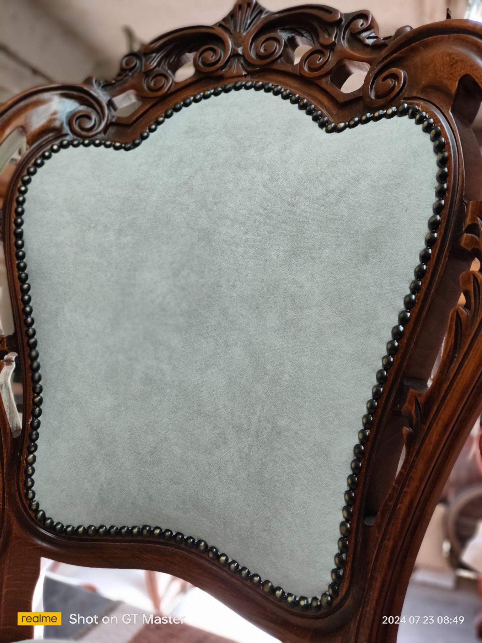 Detale odnowionego krzesła w stylu vintage: drewniana rama z rzeźbieniami i tapicerowane, jasnoszare siedzisko z ozdobnymi ćwiekami.