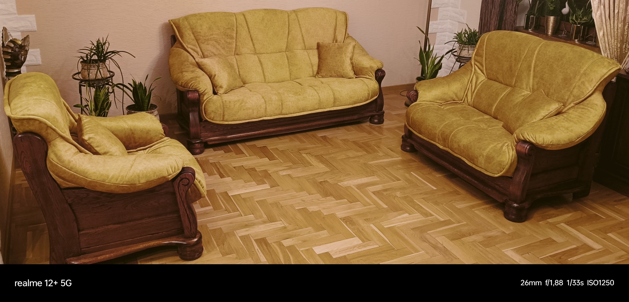 Komplet wypoczynkowy w odcieniach żółci i brązu: sofa dwuosobowa, fotel i krzesło, ustawione na parkiecie w jodełkę, uzupełnione roślinami doniczkowymi i dekoracyjną figurką.