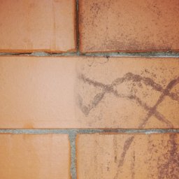 Czyściek - Fragment ściany z czerwonej cegły z widocznym śladem po czyszczeniu, gdzie jedna część cegły jest czysta, a druga zabrudzona zarysowaniami.