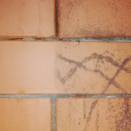 Fragment ściany z czerwonej cegły z widocznym śladem po czyszczeniu, gdzie jedna część cegły jest czysta, a druga zabrudzona zarysowaniami.