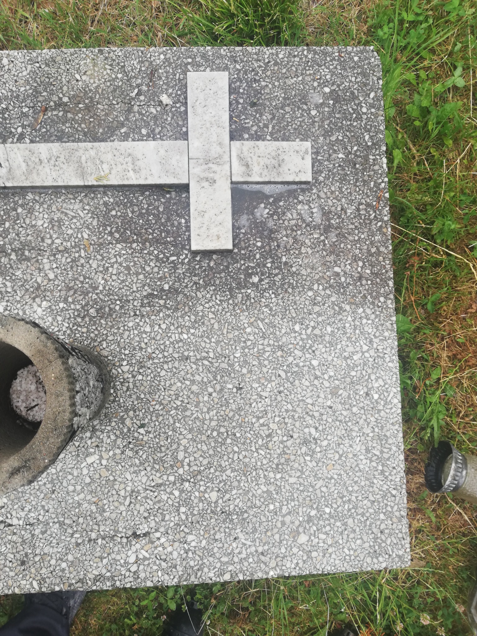 Płyta nagrobna z krzyżem z jasnego kamienia na tle trawy, z widocznym betonowym wazonem wypełnionym białymi kamykami i fragmentem butelki.