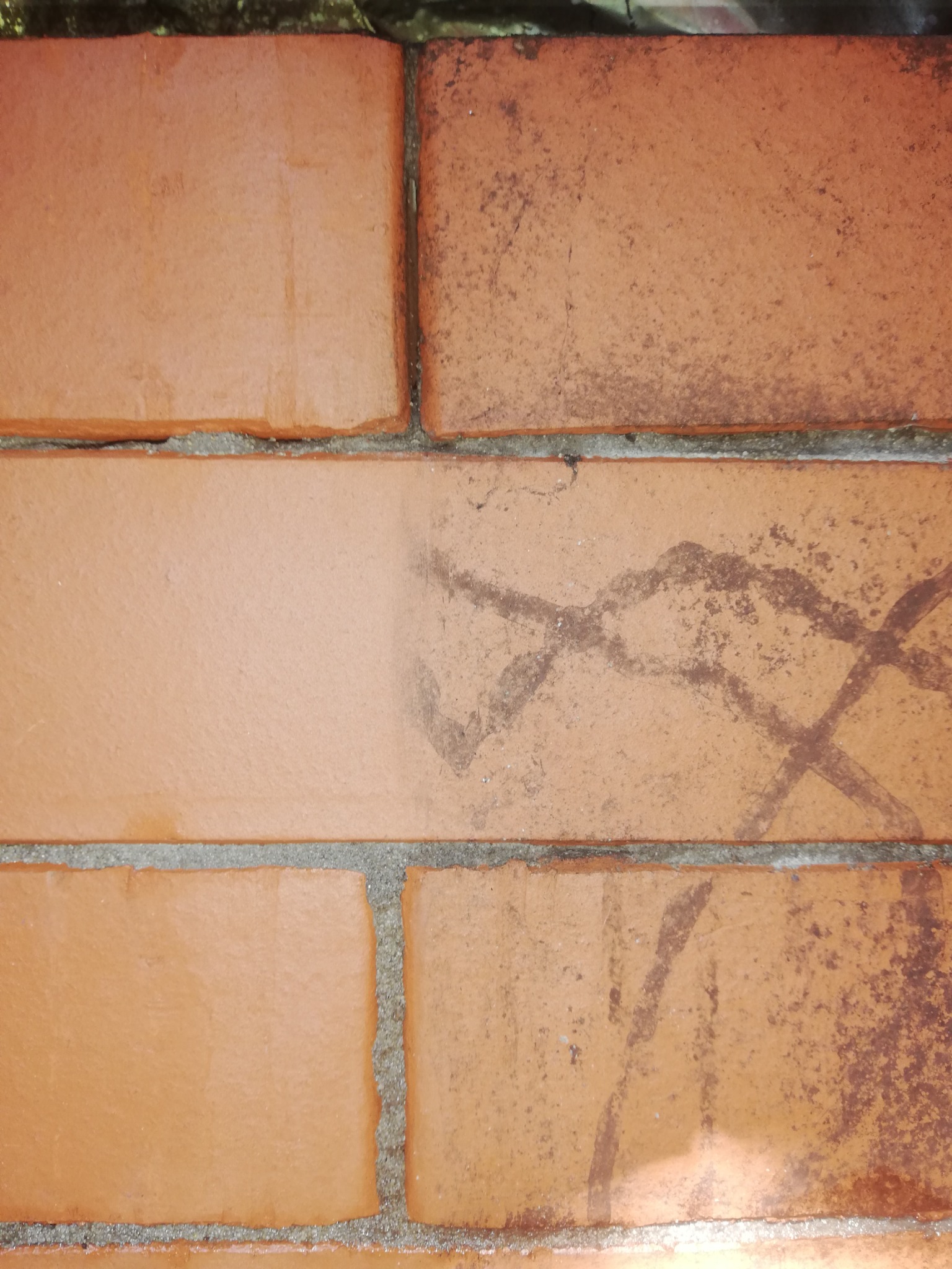 Fragment ściany z czerwonej cegły z widocznym śladem po czyszczeniu, gdzie jedna część cegły jest czysta, a druga zabrudzona zarysowaniami.