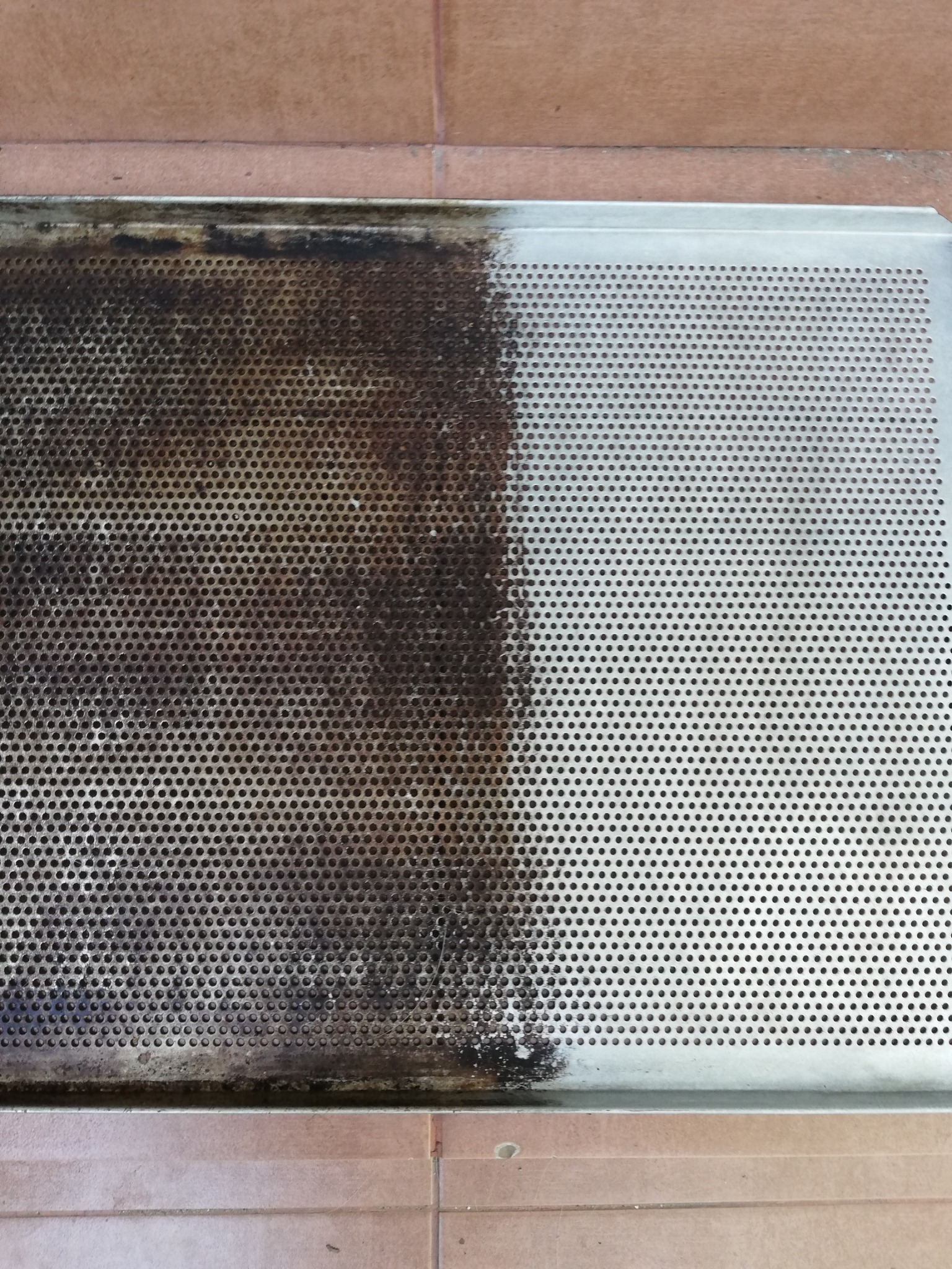 Metalowa, perforowana taca, w połowie wyczyszczona, z widocznym brudem i zabrudzeniami po lewej stronie i czystą powierzchnią po prawej stronie, leżąca na podłodze z płytek.
