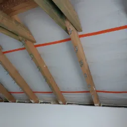 Poddasze w trakcie ocieplania: drewniane krokwie z metalowymi łącznikami, folia paroizolacyjna z pomarańczowym paskiem, szczeliny wypełnione pianką montażową.