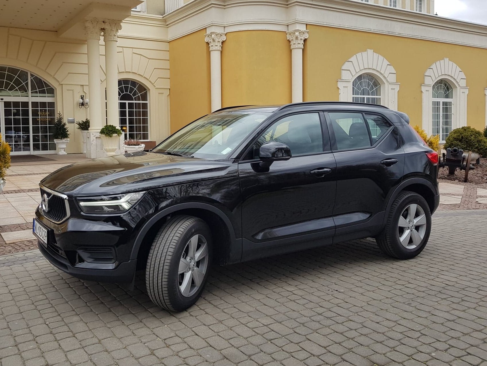 Czarny samochód Volvo XC40 zaparkowany przed budynkiem w stylu pałacowym z kolumnami i dekoracyjnymi oknami.