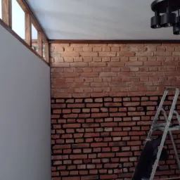 Częściowo pomalowana ceglana ściana w pomieszczeniu, górna część w kolorze ceglastym, dolna w kolorze czarnym, drabina oparta o ścianę, widoczny fragment białej ściany i drewnianej ramy okiennej...