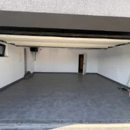 Wykończony garaż z szarą podłogą z płytek, białe ściany, widoczne narzędzia i materiały budowlane w rogu, odpływ liniowy przy wjeździe.