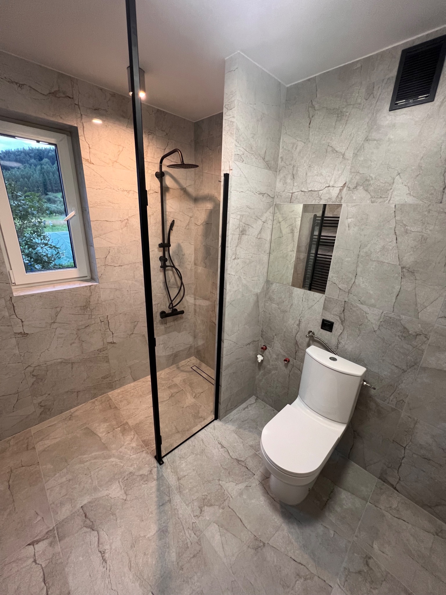 Nowoczesna łazienka w Krakowie z kabiną prysznicową typu walk-in z czarnymi profilami, marmurkowymi płytkami na ścianach i podłodze, oraz toaletą w minimalistycznym stylu. Za oknem widok na zieleń.