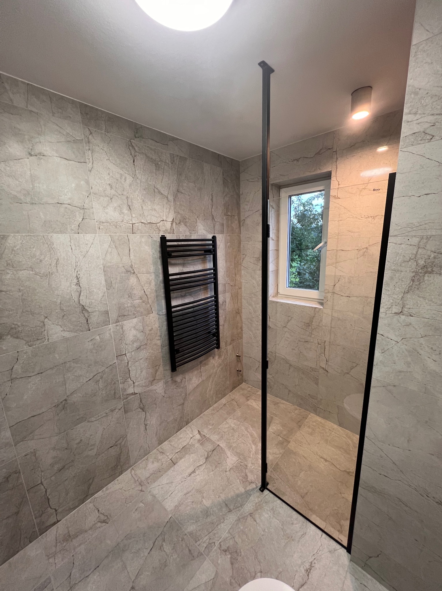 Wykończona łazienka w szarościach z czarną kabiną prysznicową typu walk-in i grzejnikiem drabinkowym, widok na okno z zielenią na zewnątrz.