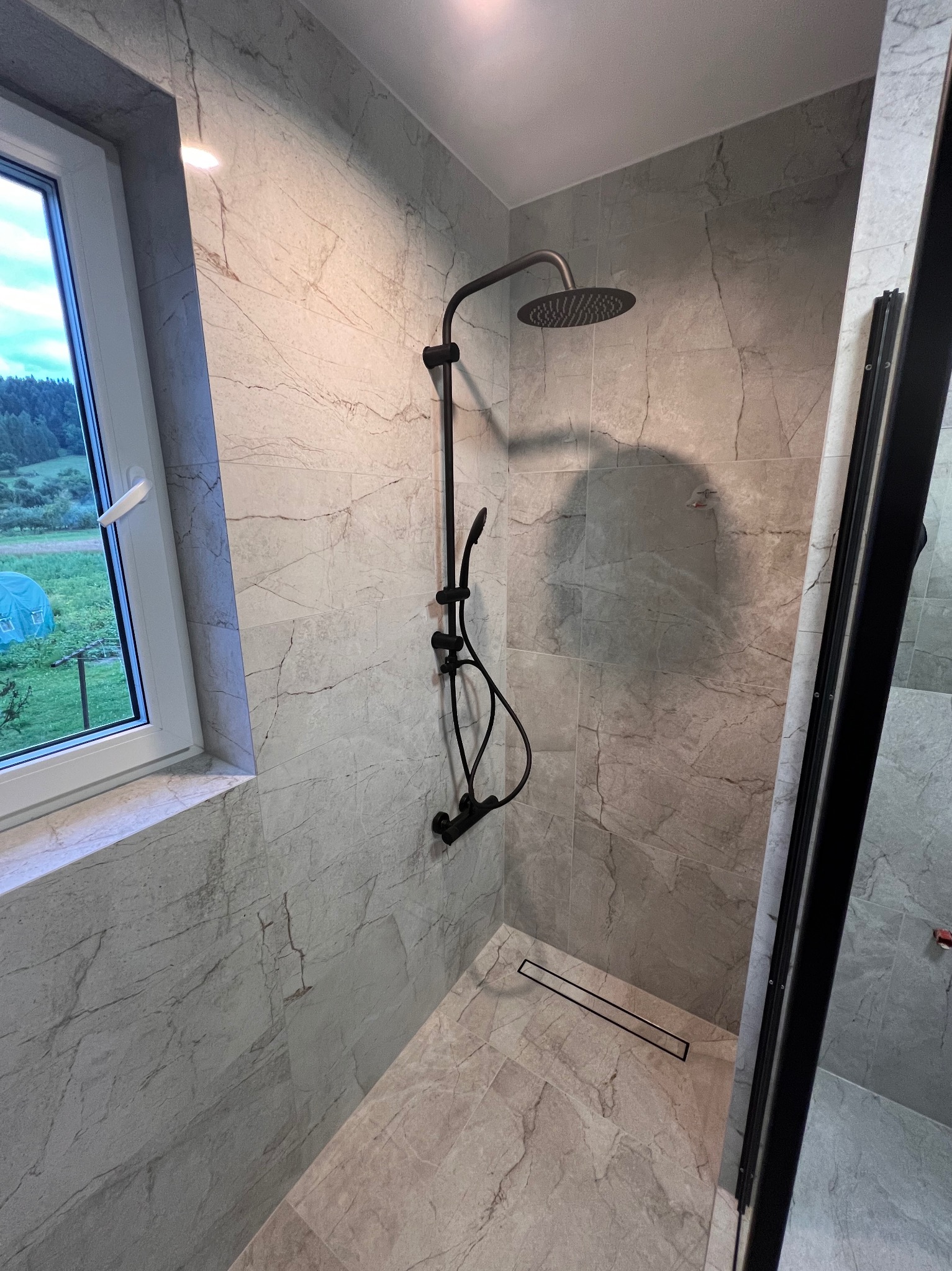 Nowoczesna kabina prysznicowa z czarną armaturą i odpływem liniowym, wykończona płytkami imitującymi marmur, widok przez okno na zielony krajobraz.