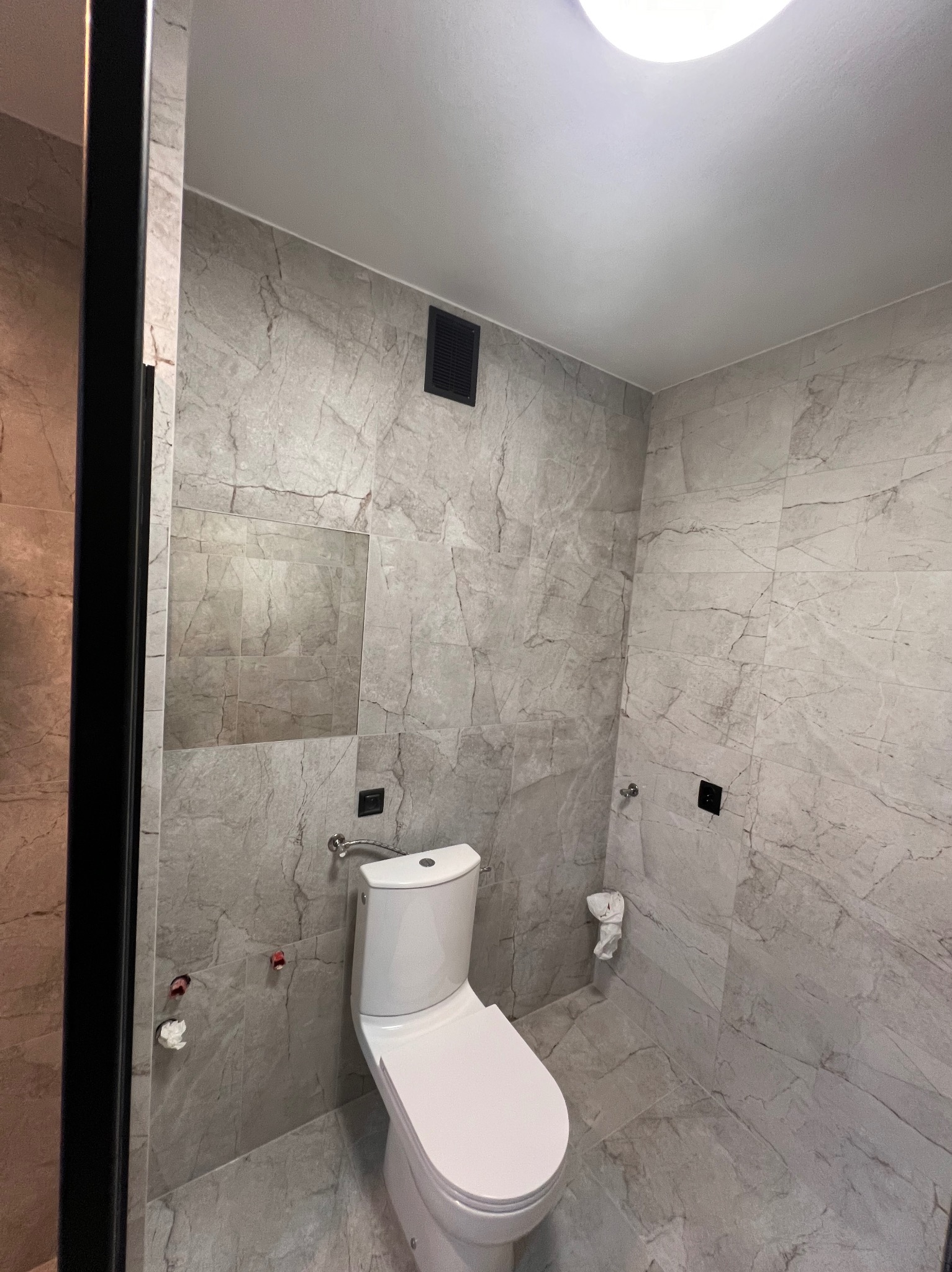 Wykończona łazienka z toaletą, szarymi płytkami imitującymi marmur na ścianach i podłodze, czarnymi akcentami w postaci kratki wentylacyjnej i ramy kabiny prysznicowej.