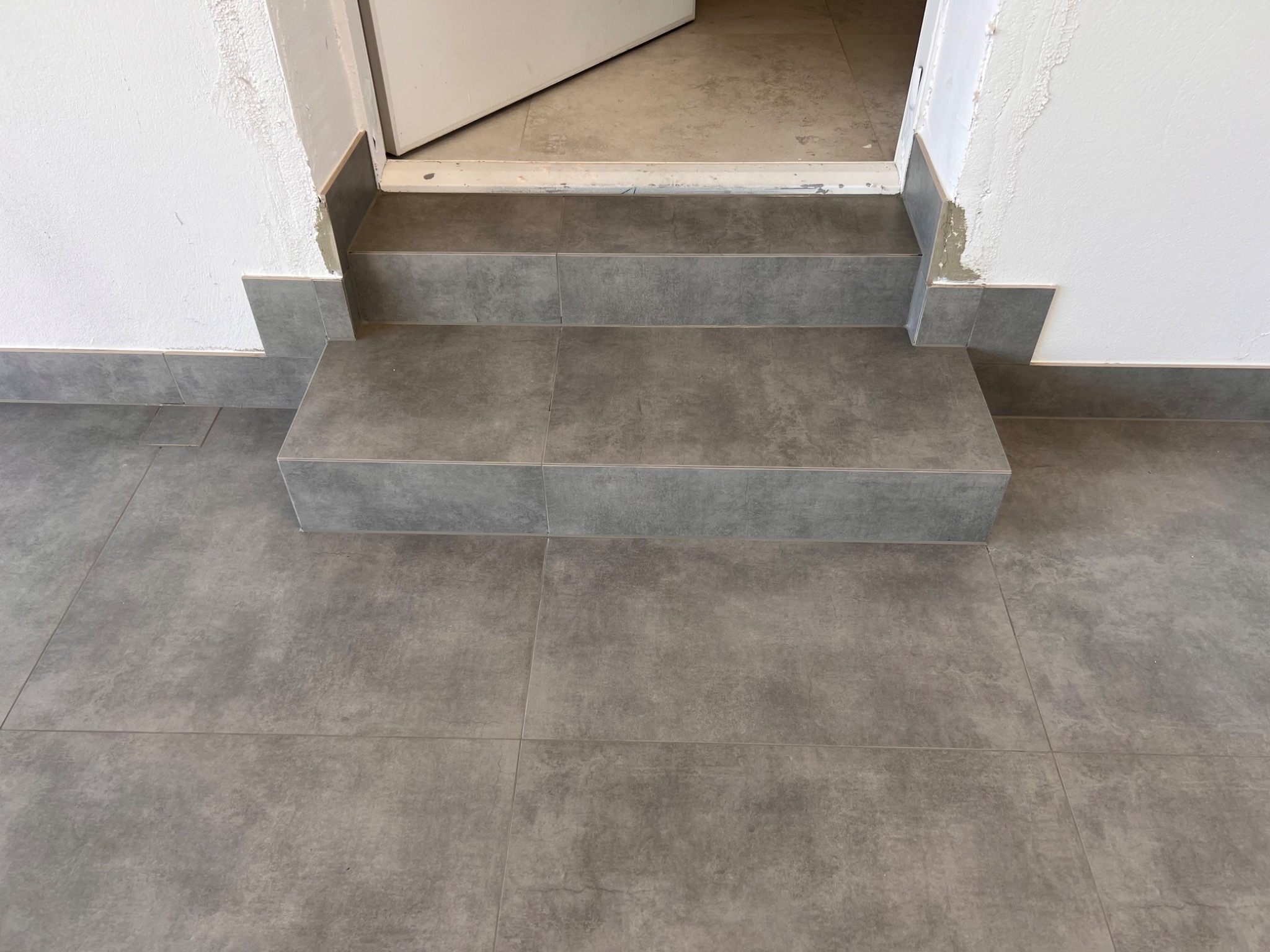 Wykończone schody wejściowe do pomieszczenia z szarych, kwadratowych płytek ceramicznych, z widocznymi niedoskonałościami wykończenia przy ścianie i ościeżnicy drzwiowej.